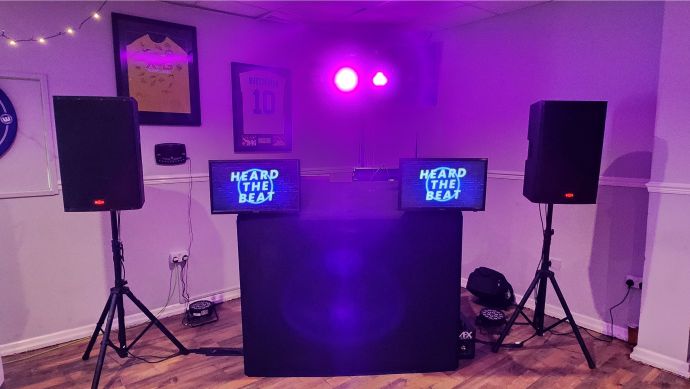 Heard The Beat DJ booth setup with dual display screens, lighting effects, and professional speakers in a venue with wood flooring and football memorabilia