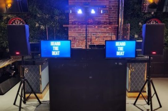 Black DJ booth setup with dual “Heard The Beat” display screens, speaker stands, and LED lighting in a stylish venue with brick and greenery backdrop.
