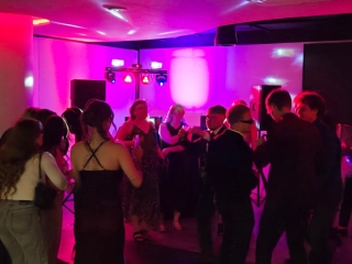 Guests dancing in a dark room with vibrant pink and purple DJ lighting and sound equipment creating a lively party atmosphere.