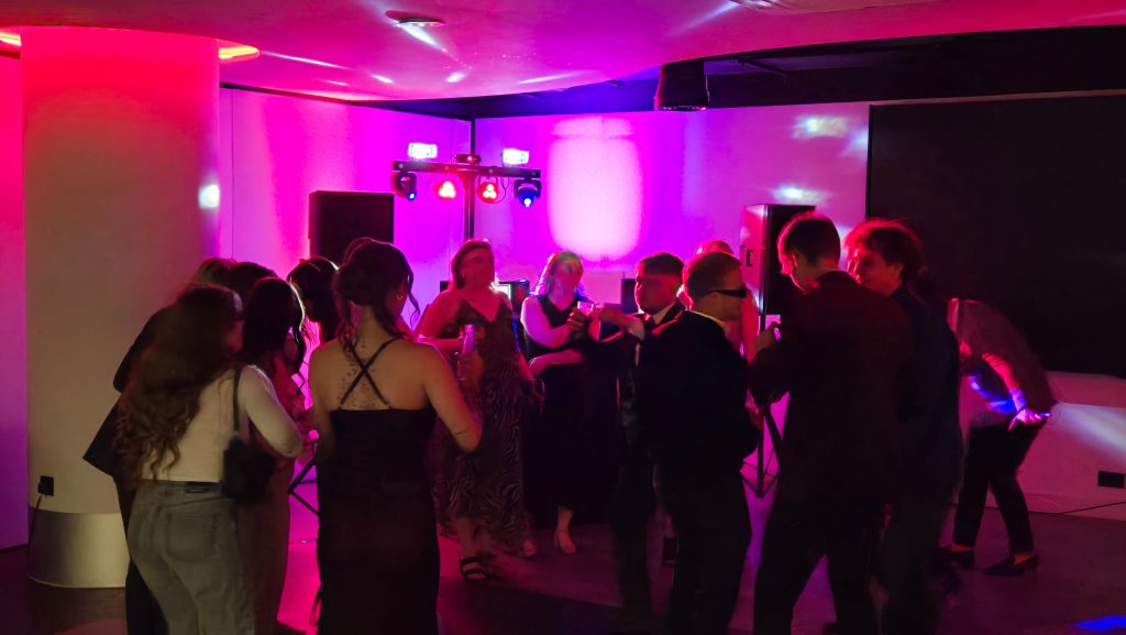 Guests dancing in a dark room with vibrant pink and purple DJ lighting and sound equipment creating a lively party atmosphere.
