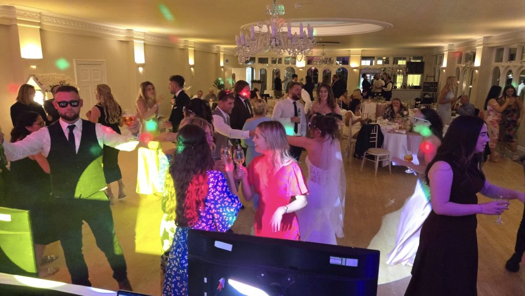Wedding guests dancing under colorful DJ lighting at an evening reception, with a lively atmosphere and bride joining the celebration.