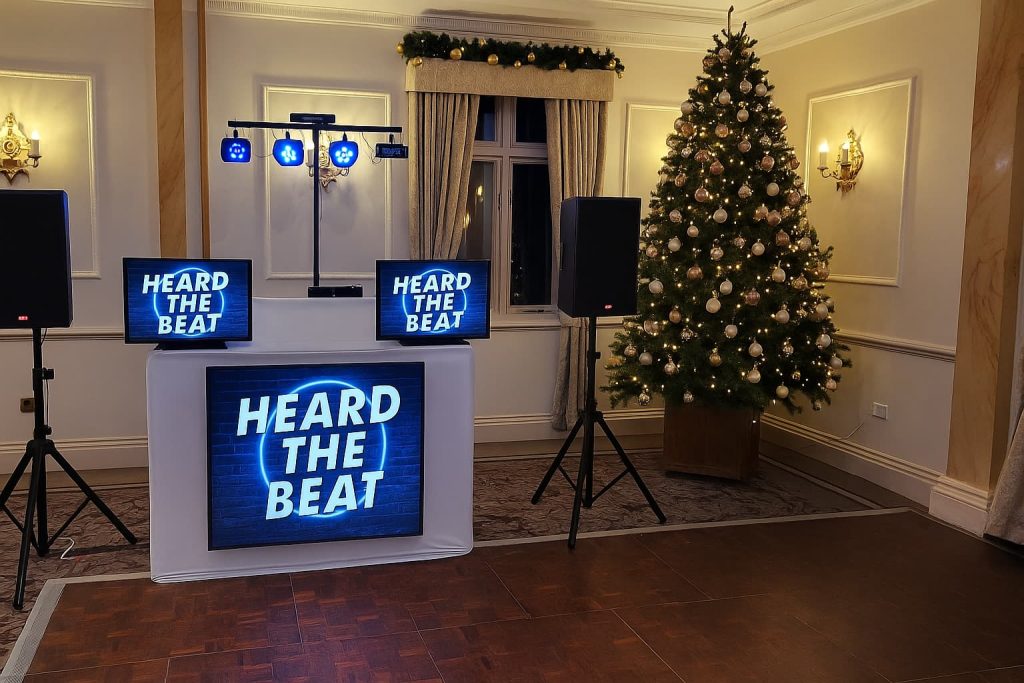 Heard The Beat DJ setup in a decorated function room with a large Christmas tree, two side screens and a central screen on the booth all displaying the Heard The Beat logo.