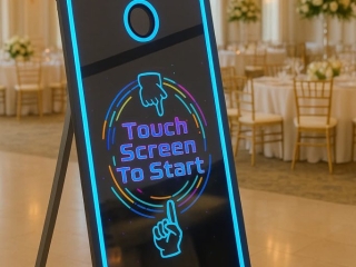 A sleek, interactive mirror photo booth with neon blue accents stands in an elegant wedding venue featuring round tables, gold chairs, floral centerpieces, and chandeliers. The booth displays a colorful "Touch Screen To Start" prompt.