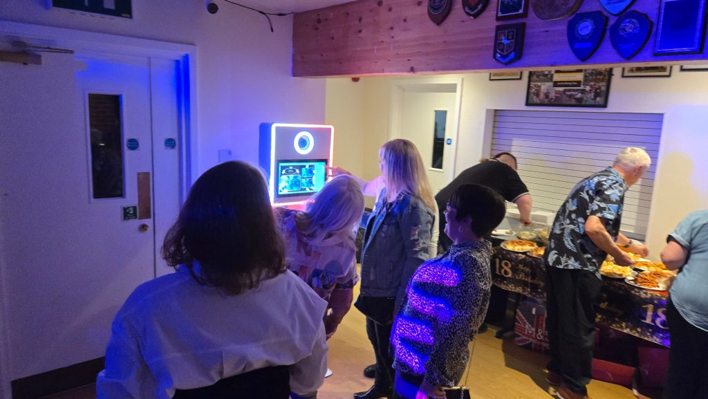 Guests gather around a compact LED photo booth at an indoor party, smiling and interacting with the touch screen while others enjoy food from a buffet table in the background.