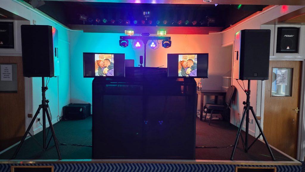 Professional DJ setup on a stage featuring two large speakers on tripods, a black DJ booth, dual display screens showing a stylized image of a couple, and a colourful lighting rig with uplighting and effect lights creating a vibrant party atmosphere.