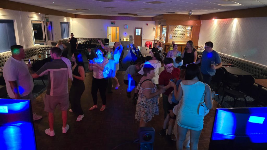 A lively group of party guests dancing and mingling on a wooden dance floor in a warmly lit function room, with blue lighting effects adding a festive atmosphere. People are enjoying drinks, taking photos, and chatting, with tables and seating arranged around the edges of the room.