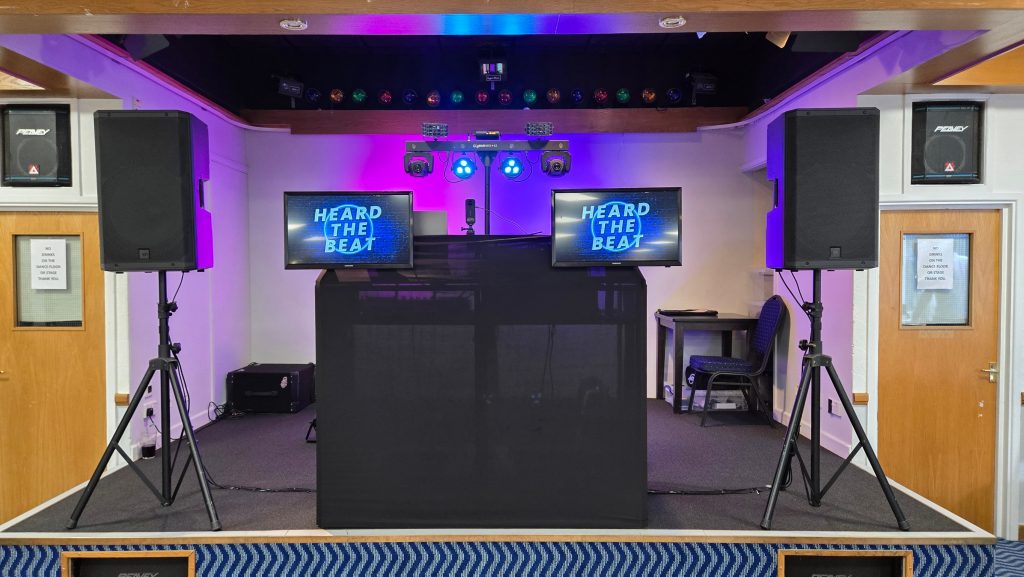 Professional DJ stage setup featuring a black DJ booth, two large speakers on tripods, dual screens displaying the “Heard The Beat” logo, and an overhead lighting rig with purple uplighting and LED effects, ready for an event. The setup is positioned on a raised stage in a function room.