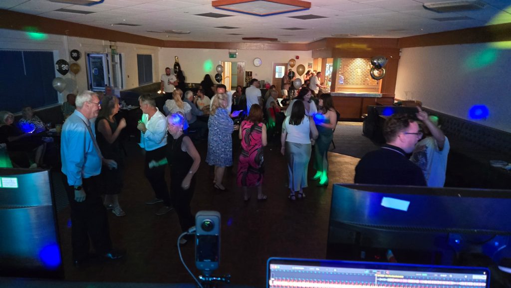 Party guests of various ages dancing and enjoying themselves in a function room decorated with 30th birthday balloons. The photo is taken from behind the DJ booth, showing the DJ’s equipment and monitors in the foreground, with colourful lights casting effects across the lively crowd and surrounding seating area