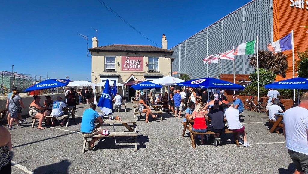 Crowds enjoying a sunny outdoor event at The Ship & Castle pub in Portsmouth, with people seated at wooden benches under Foster’s-branded umbrellas, surrounded by flags and a self-storage building in the background.