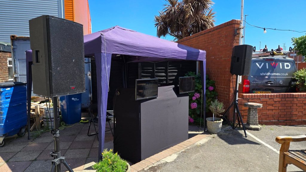 Outdoor DJ setup under a purple gazebo with large black speakers on stands, dual screen DJ booth with "Heard The Beat" branding, located in a sunny courtyard next to a Vivid Homes van and surrounded by brick walls, plants, and urban decor.