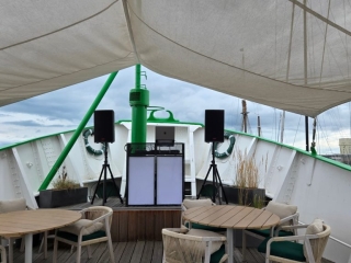A DJ booth with speakers set up on the front deck of a boat under a canopy, surrounded by empty tables and chairs, with a cloudy sky and marina in the background.