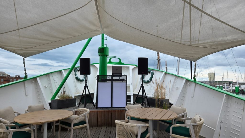 A DJ booth with speakers set up on the front deck of a boat under a canopy, surrounded by empty tables and chairs, with a cloudy sky and marina in the background.