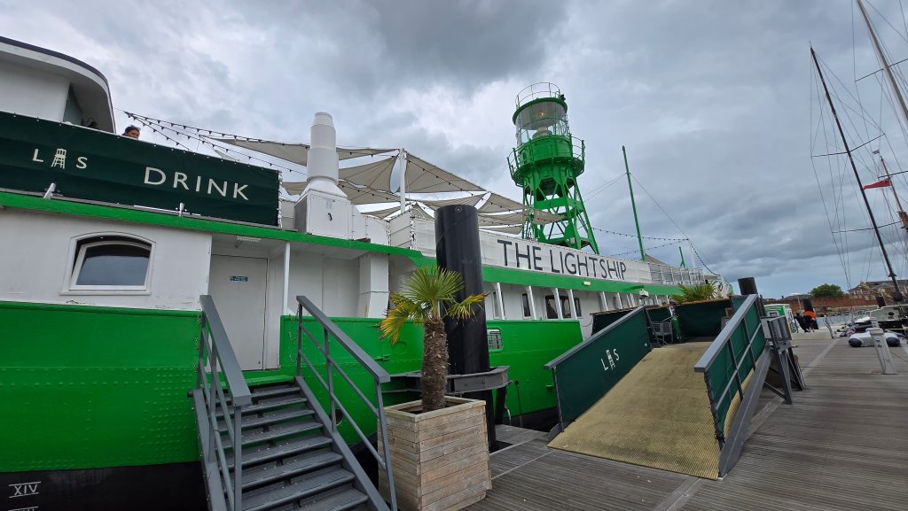 A view of The Lightship, a vibrant green and white docked ship converted into a venue, with entrance stairs and a ramp, outdoor canopy area, and a green lighthouse structure on top, under a cloudy sky at the marina.