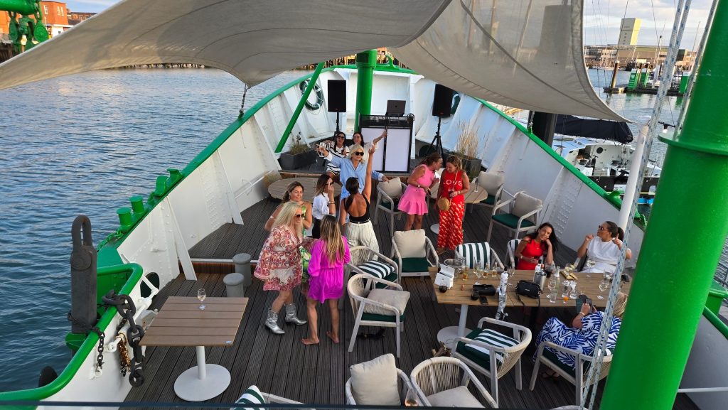 A group of women gathered on the front deck of a boat during the day, some dancing and others seated around tables with drinks, under a large canopy and surrounded by calm harbor waters.