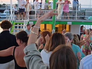 A lively group of people dancing and enjoying a party on the deck of a boat at sunset, with guests on both upper and lower levels, festive lighting, and a relaxed summer atmosphere.