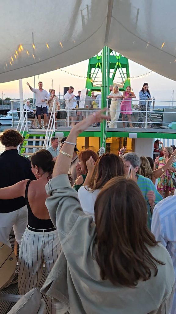A lively group of people dancing and enjoying a party on the deck of a boat at sunset, with guests on both upper and lower levels, festive lighting, and a relaxed summer atmosphere.