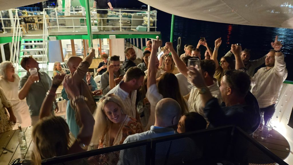 A lively nighttime party on a boat with a large group of guests dancing and raising their hands in celebration, surrounded by soft lighting and reflections on the water in the background.