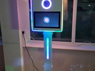 A sleek LED photo booth with a glowing light ring and screen, topped with a silver umbrella light, set up indoors at an event.