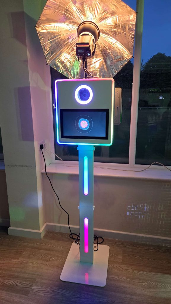 A sleek LED photo booth with a glowing light ring and screen, topped with a silver umbrella light, set up indoors at an event.