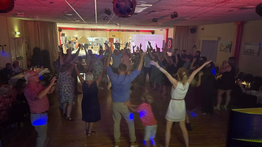Party guests at The Royal Naval Association Gosport dancing with their arms raised during a group dance led by the DJ.