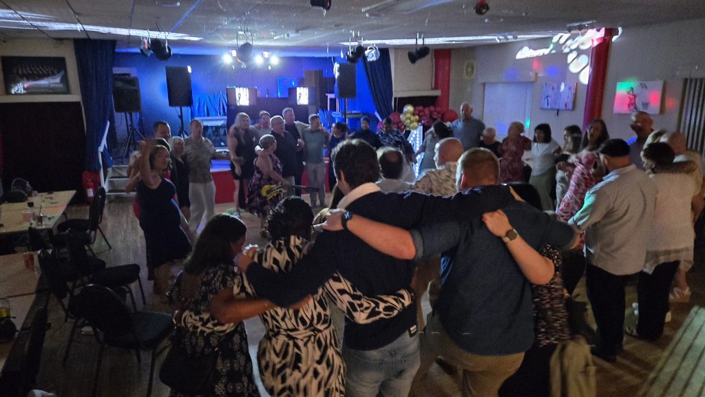 Guests arm in arm enjoying a lively circle dance at The Royal Naval Association Gosport with DJ music and lighting.