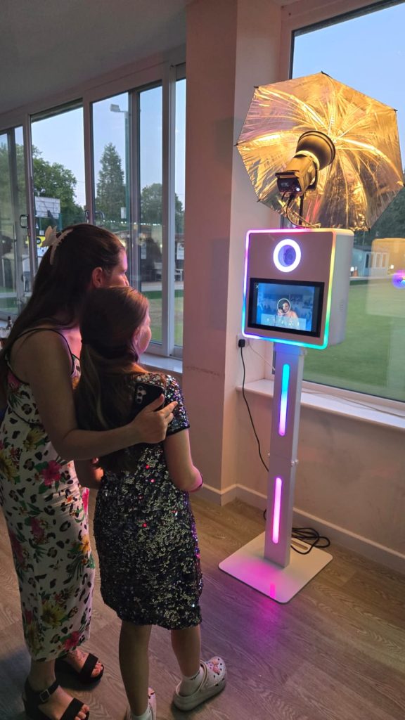 A woman and young girl posing together in front of an LED photo booth at an indoor evening event.