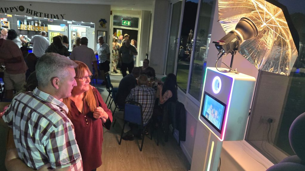 A couple smiling at an LED photo booth during a lively indoor birthday celebration with guests in the background.