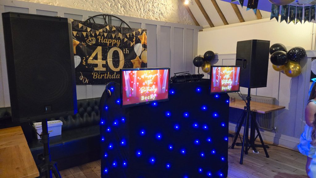 Professional DJ booth with blue LED lights, speakers, and personalised screens for a 40th birthday celebration by Heard The Beat.