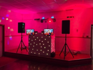Red and pink uplighting DJ stage setup with starlit booth, speakers, and dual video screens for party entertainment.