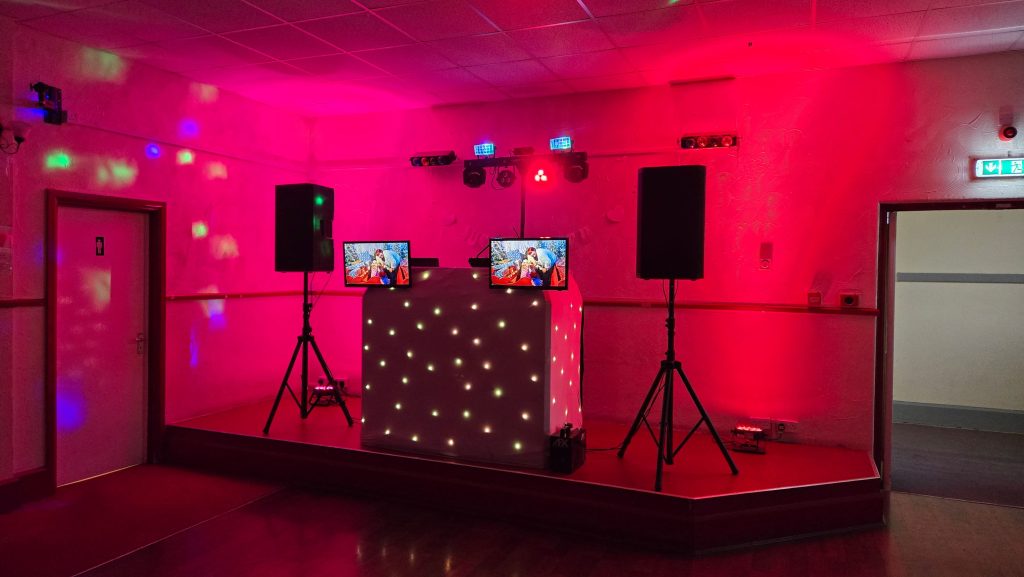 Red and pink uplighting DJ stage setup with starlit booth, speakers, and dual video screens for party entertainment.